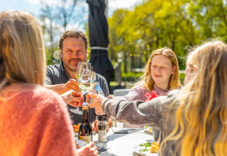 Familie nyder et måltid og skåler sammen udendørs på en solrig dag i et feriepark-glampingområde.