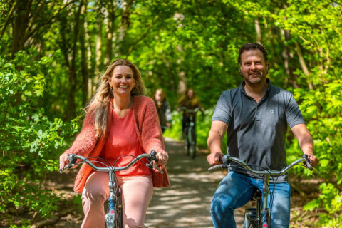 Adultos sonrientes pasean en bicicleta por un sendero forestal en un parque vacacional con glamping.