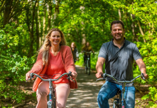 Deux adultes souriants font du vélo sur un chemin boisé d’un parc de vacances proposant du glamping.