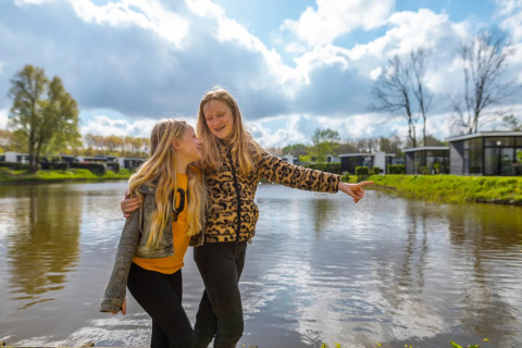 Twee lachende meisjes knuffelen bij een vijver op een vakantiepark met glamping-accommodaties.