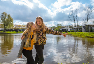 Twee lachende meisjes knuffelen bij een vijver op een vakantiepark met glamping-accommodaties.