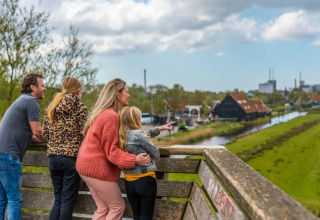 Gezin bewondert het uitzicht vanaf een terras in een vakantiepark met glamping, groene velden en water.