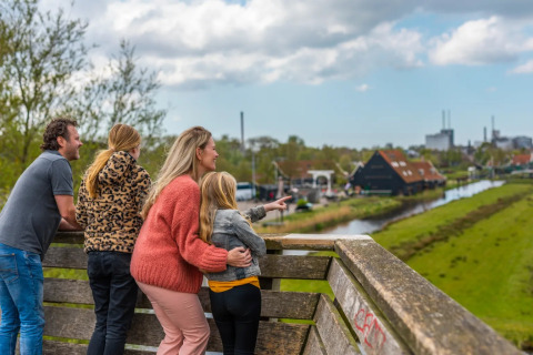 Famille admirant la vue depuis un balcon d’un parc de vacances avec glamping, champs verts et canal.