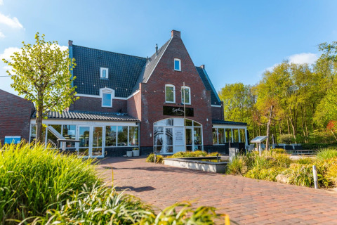 Foto van een vakantiepark met glamping, bakstenen gebouw, terras, groene beplanting en blauwe lucht.