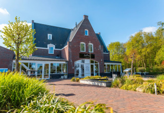 Foto van een vakantiepark met glamping, bakstenen gebouw, terras, groene beplanting en blauwe lucht.