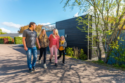 Familia de cuatro pasea frente a una cabaña glamping moderna en un parque de vacaciones bajo cielo soleado.