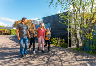 Familie på fire går foran et moderne glampinghus i en feriepark på en solrig dag med blå himmel.