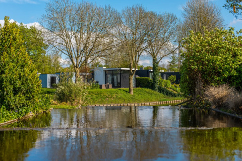 Modern glamping lodges by a calm pond surrounded by lush greenery in a holiday park under blue skies.