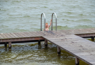 Barn klatrer op ad en badestige ved en træbro over søen i en feriepark med glampingfaciliteter.