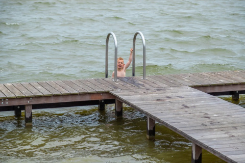 Child climbing a swim ladder attached to a wooden pier over a lake at a holiday park with glamping.