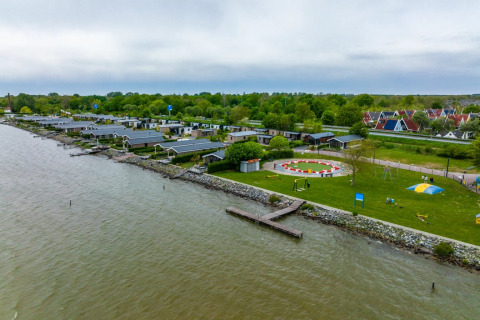 Vista aérea de un parque vacacional con alojamientos glamping junto al agua, áreas verdes y parque infantil.