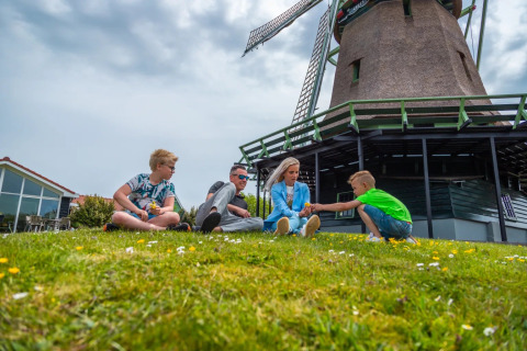 Family relaxing on the grass in front of a Dutch windmill at a holiday park offering glamping stays.