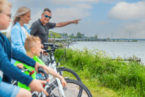 Familia paseando en bicicleta junto a un lago con tiendas glamping y aerogeneradores en un parque vacacional.