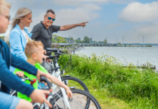 Familie på cykeltur ved en sø med glampinghytter og vindmøller i baggrunden i en feriepark.