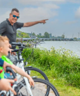 Familia paseando en bicicleta junto a un lago con tiendas glamping y aerogeneradores en un parque vacacional.