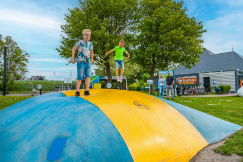 Dos niños juegan en una gran almohada inflable en un parque vacacional con glamping y áreas verdes.