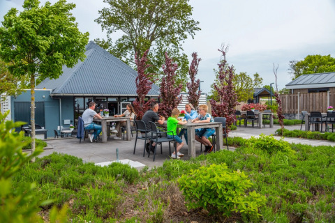 Udendørs spiseområde på en feriepark med glamping, familier nyder måltider omgivet af grønne planter.