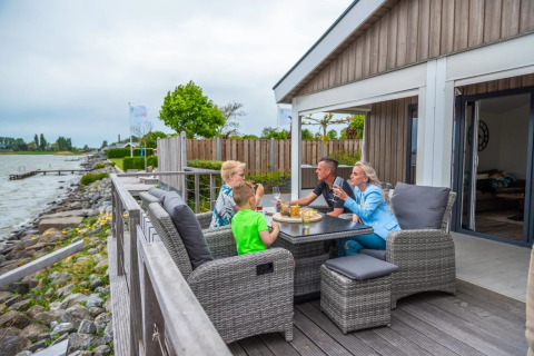 Familia disfrutando de una comida en la terraza junto al agua en un parque de vacaciones glamping.