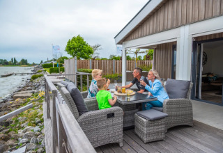 Familie nyder et måltid på terrassen ved vandet i et glamping-ophold på en feriepark.