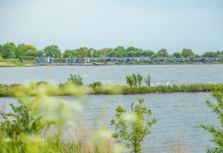 Udsigt over en sø med glamping-hytter og frodig natur i en feriepark på en rolig, overskyet dag.