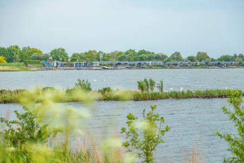 Vista de un lago con cabañas glamping y vegetación en un parque vacacional en un tranquilo día nublado.
