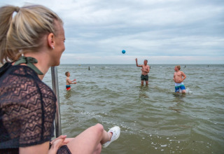 Vrouw kijkt toe terwijl gezin met bal speelt in zee bij een vakantiepark met glampingaccommodaties.