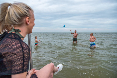 Mujer observa a una familia jugando a la pelota en el mar en un parque vacacional con alojamientos glamping.
