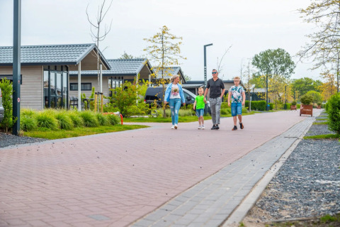Family walks along a pathway between modern glamping cabins at a holiday park with green gardens around.