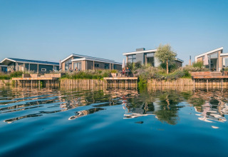 Billede af moderne glamping-hytter ved vandet i en feriepark med spejlinger i det rolige vand.