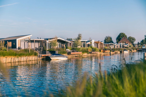 Cabanes de glamping modernes au bord d’un canal dans un parc de vacances, entourées de verdure et d’eau calme.