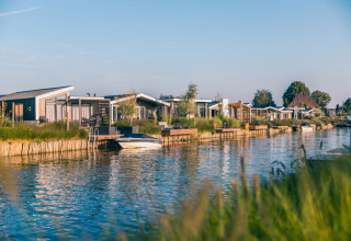 Moderne glamping-hytter ved en kanal på en feriested, omgivet af grønt og spejlblankt vand under blå himmel.