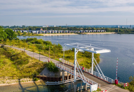 Blick auf eine Brücke am See mit modernen Glamping-Unterkünften im Hintergrund eines Ferienparks.