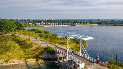 Uitzicht op een brug bij een meer met moderne glamping-accommodaties op de achtergrond in een vakantiepark.