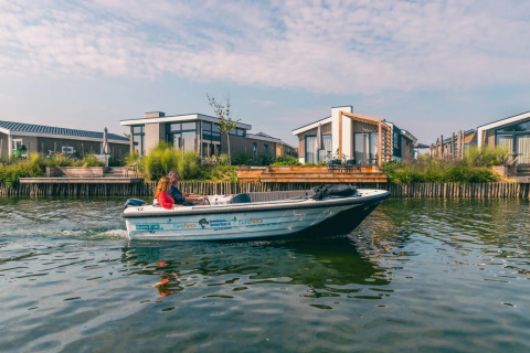 Un bote navega cerca de modernas cabañas de glamping en un parque vacacional junto al agua.