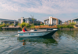 En familie sejler i en lille båd foran moderne glamping-hytter ved vandet i en feriepark.