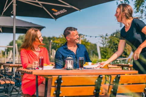Couple profitant d’un repas en terrasse dans un parc de vacances, servi par une serveuse souriante.