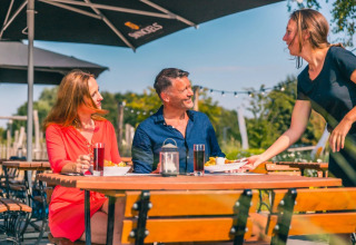 Par nyder mad og drikke på en udendørs café i solskin, serveret af en smilende kvinde på feriepark.