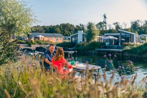 Koppel eet buiten aan het water in een vakantiepark met moderne glampingverblijven en groene omgeving.
