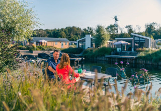 Koppel eet buiten aan het water in een vakantiepark met moderne glampingverblijven en groene omgeving.