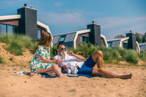 Pareja disfruta de un picnic en la playa de arena frente a cabañas glamping modernas en el parque vacacional.