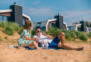 Par nyder picnic på stranden foran moderne glamping-hytter i en feriepark under solrigt vejr.