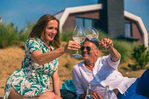 Pareja brinda con vino frente a una tienda glamping en un parque vacacional, disfrutando del sol y relajados.