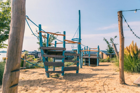 Parque infantil en arena con estructura para escalar en camping de lujo con cabañas y naturaleza.