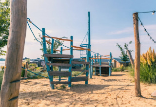 Legeplads i sandet med et klatrelegeredskab ved feriepark med glamping, hytter og grøn natur.