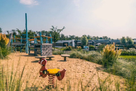 Legeplads med sand, legeredskaber og moderne glamping-hytter i baggrunden i en feriepark.
