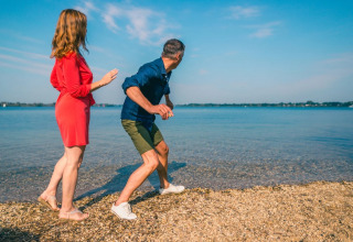 Couple enjoys lakeside at a holiday park with glamping, skipping stones under a bright blue sky.