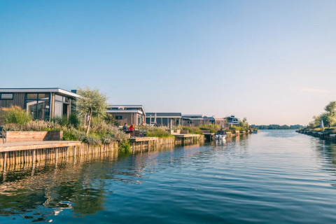 Modern glamping lodges along a calm waterfront at a holiday park, surrounded by greenery and clear skies.