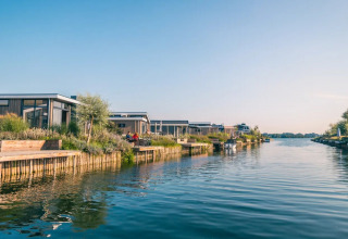 Modern glamping lodges along a calm waterfront at a holiday park, surrounded by greenery and clear skies.
