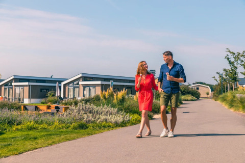 A couple enjoys ice cream while walking through a holiday park with glamping cabins on a sunny day.
