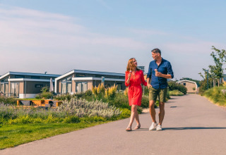 A couple enjoys ice cream while walking through a holiday park with glamping cabins on a sunny day.